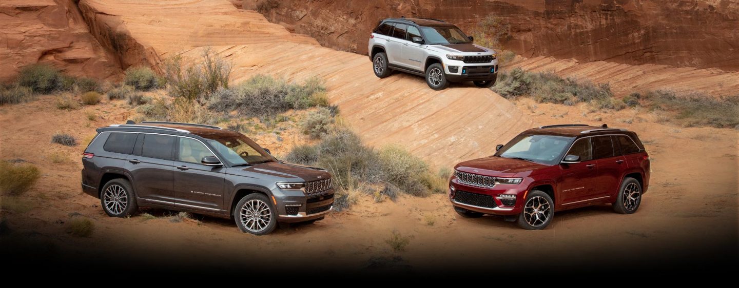 Three Jeep Grand Cherokee models, including the three-row Grand Cherokee L Summit Reserve and the Grand Cherokee 4xe, parked off-road in scrubby desert terrain.